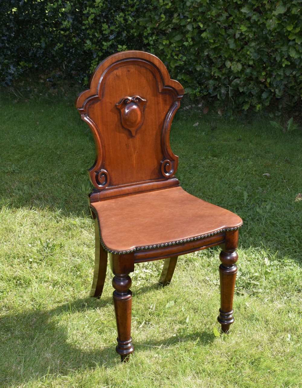 A Victorian mahogany hall chair with a carved shield-shaped backrest, stud-trimmed dark seat, turned front legs, and splayed rear legs. View XV