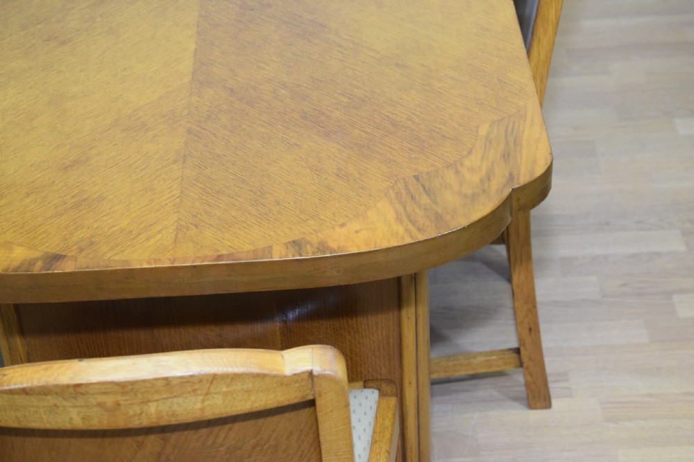An Art Deco light oak dining suite featuring a shaped table with a central pedestal surrounded by four matching upholstered chairs. View X