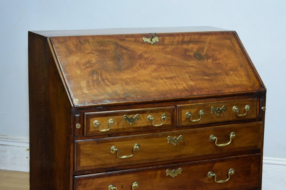 A Georgian walnut bureau featuring a sloped hinged front opening above four graduated drawers with decorative brass handles and bracket feet. View X