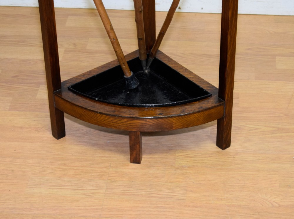 An Arts and Crafts dark oak corner stick stand featuring a curved upper front rail, straight vertical supports, and a black triangular metal drip tray. View III