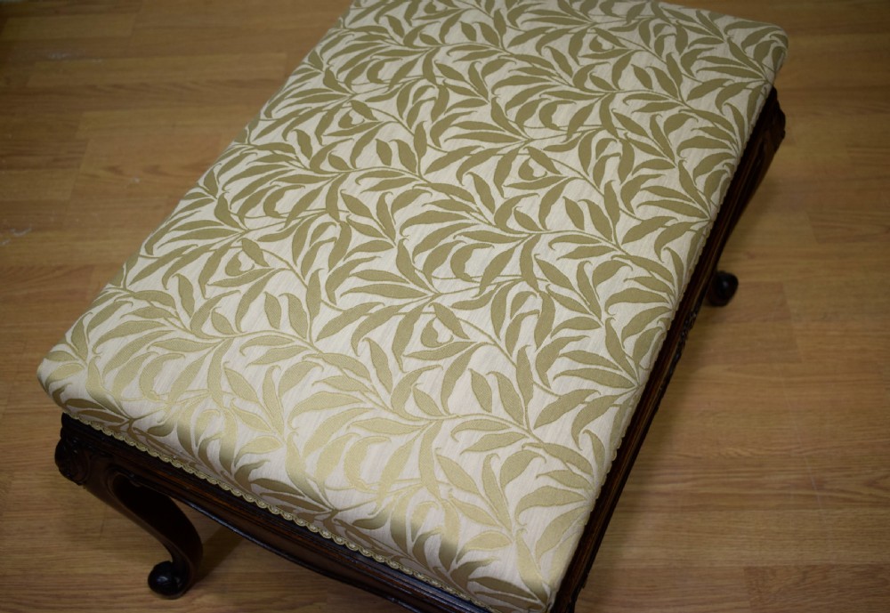An early 20th-century French stool with carved dark timber cabriole legs and a cream and gold patterned fabric upholstered top. View X