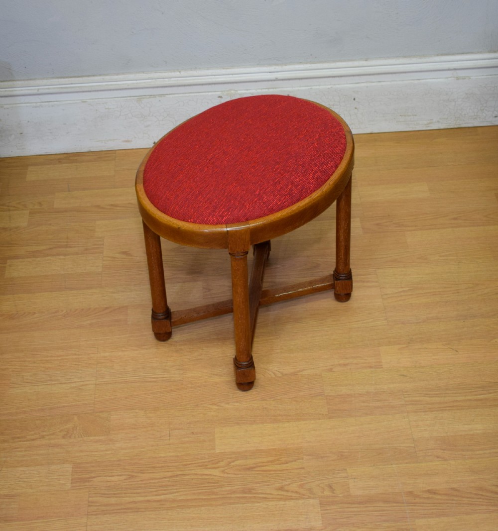 An antique Arts and Crafts oak stool featuring an oval red upholstered seat supported by four turned legs with a cross stretcher.