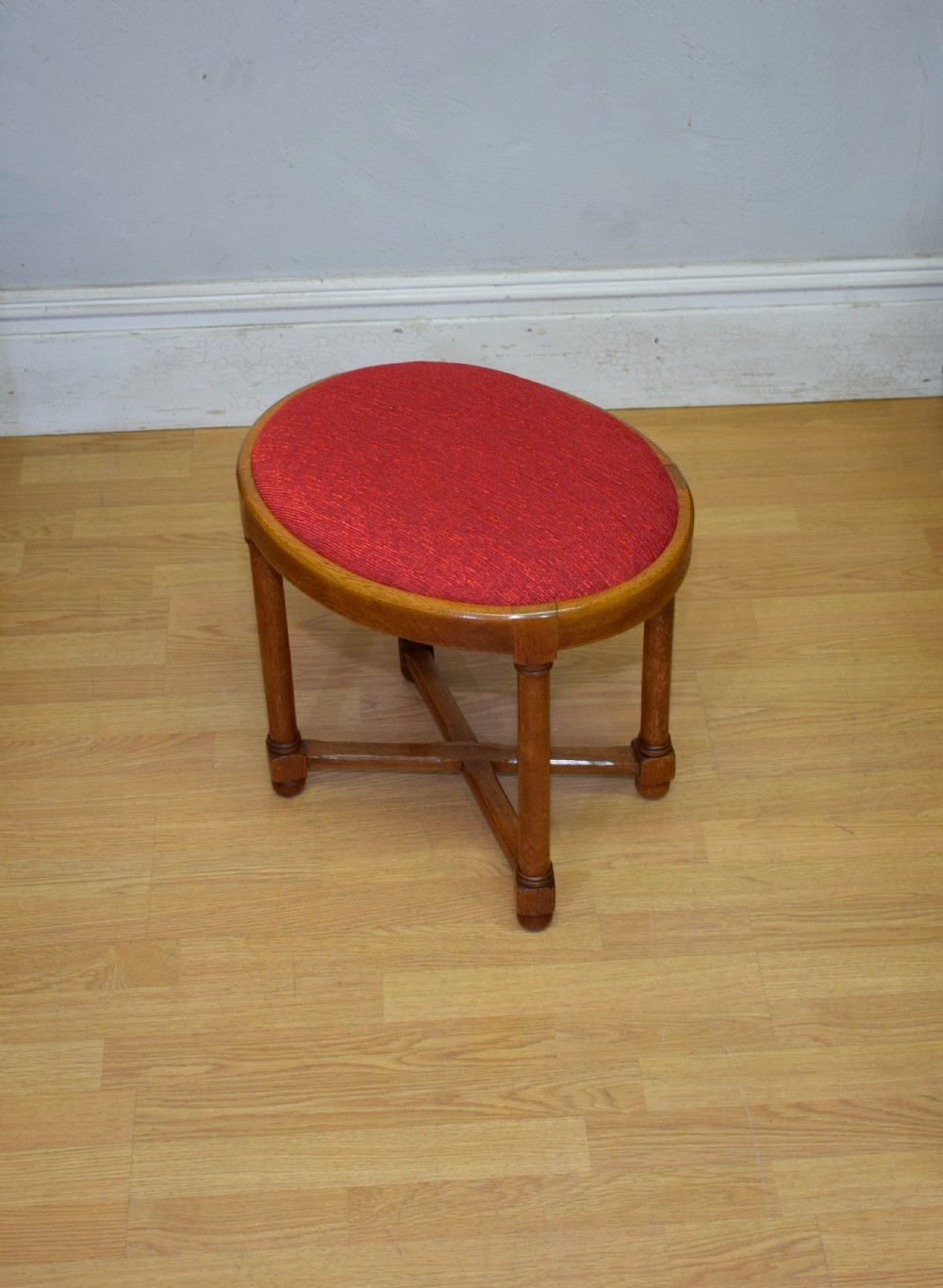 An antique Arts and Crafts oak stool featuring an oval red upholstered seat supported by four turned legs with a cross stretcher. View II