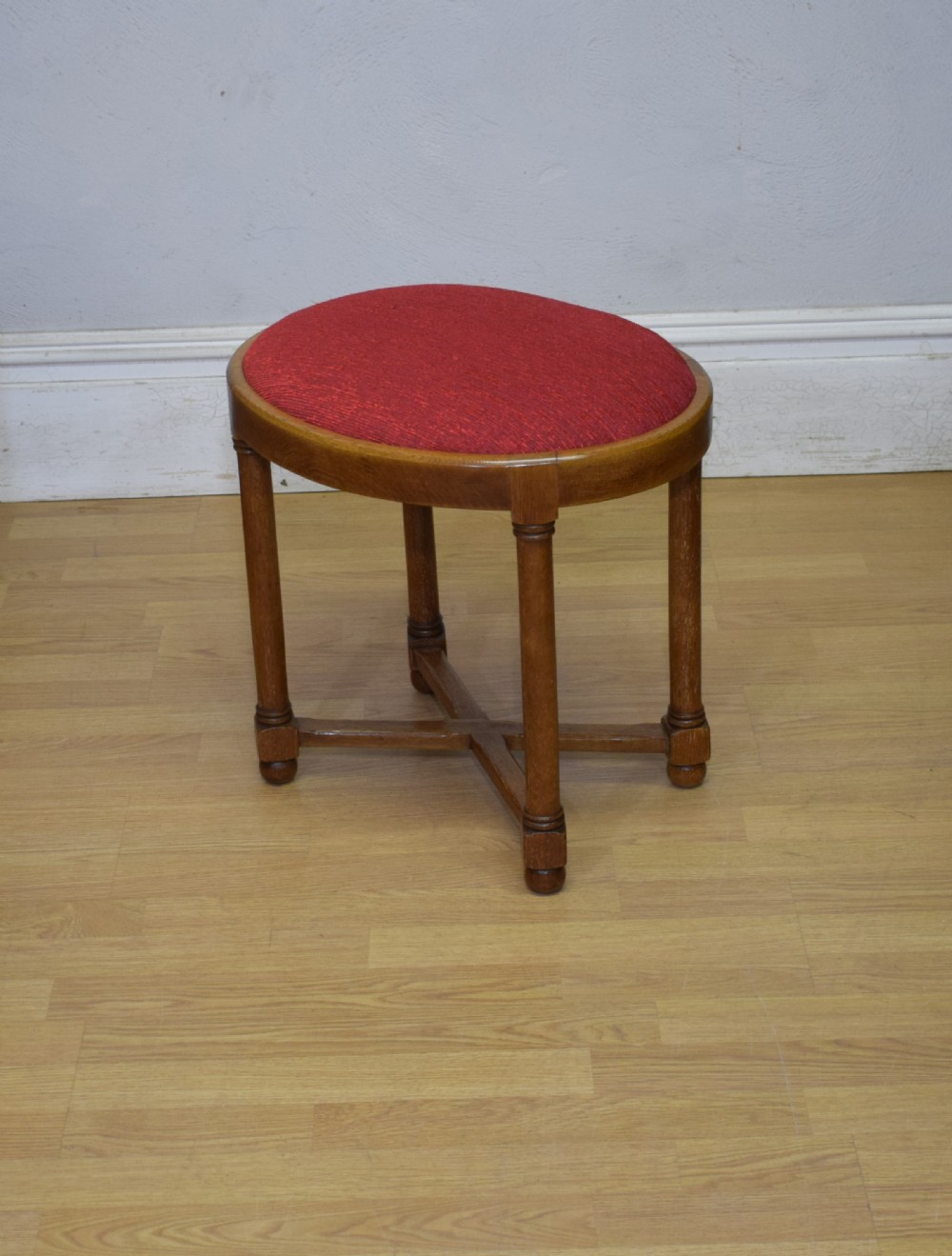 An antique Arts and Crafts oak stool featuring an oval red upholstered seat supported by four turned legs with a cross stretcher. View X