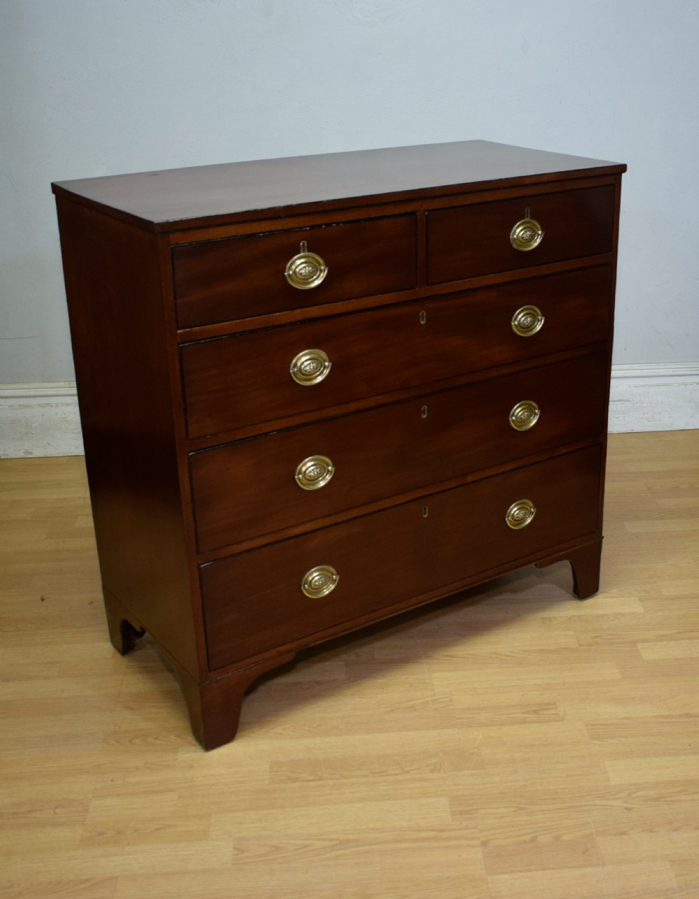 A Regency period mahogany chest of drawers featuring a two short over three long graduated drawer layout with polished brass oval handles and bracket feet. View VIII