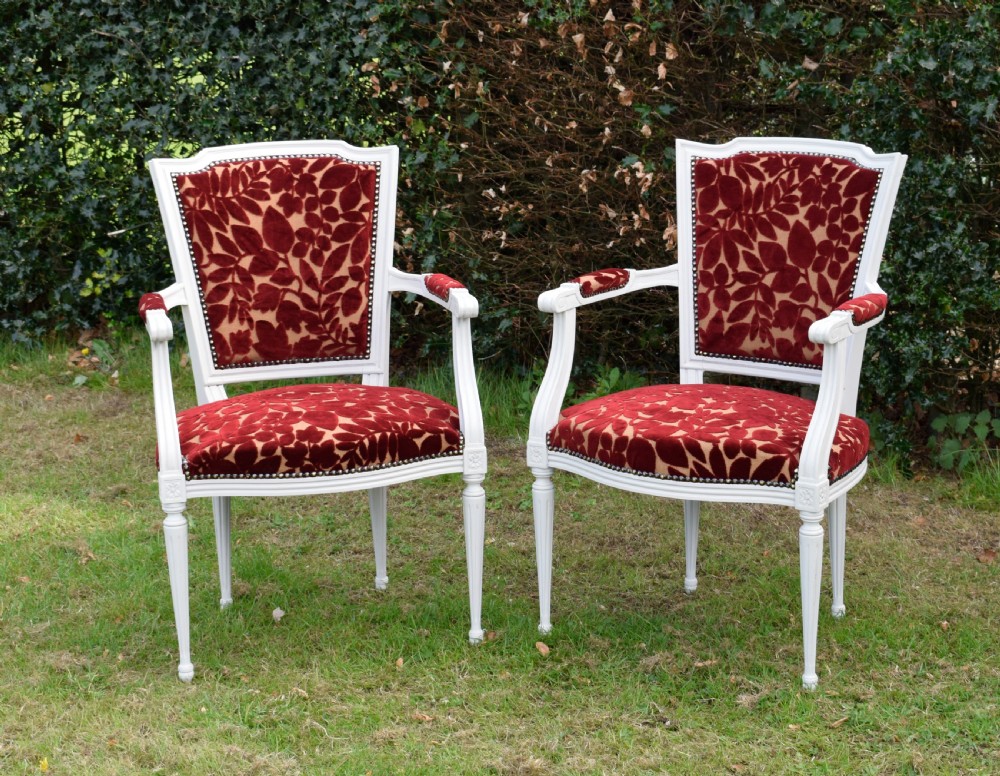 A pair of white painted open armchairs featuring fluted legs, padded armrests, and burgundy botanical cut velvet upholstery secured with brass studs.