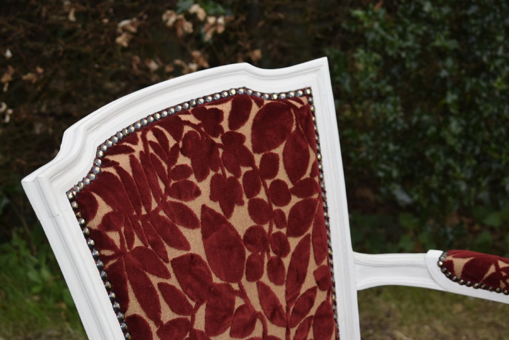 A pair of white painted open armchairs featuring fluted legs, padded armrests, and burgundy botanical cut velvet upholstery secured with brass studs. View X