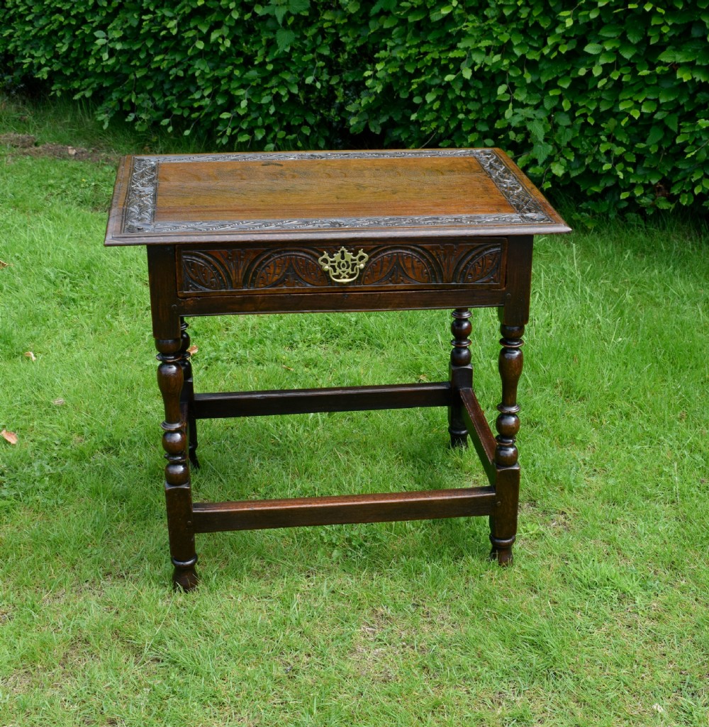 A dark oak 17th-century lowboy table featuring a carved rectangular top, a single carved drawer front with a central brass handle, turned legs, and low wooden stretchers.
