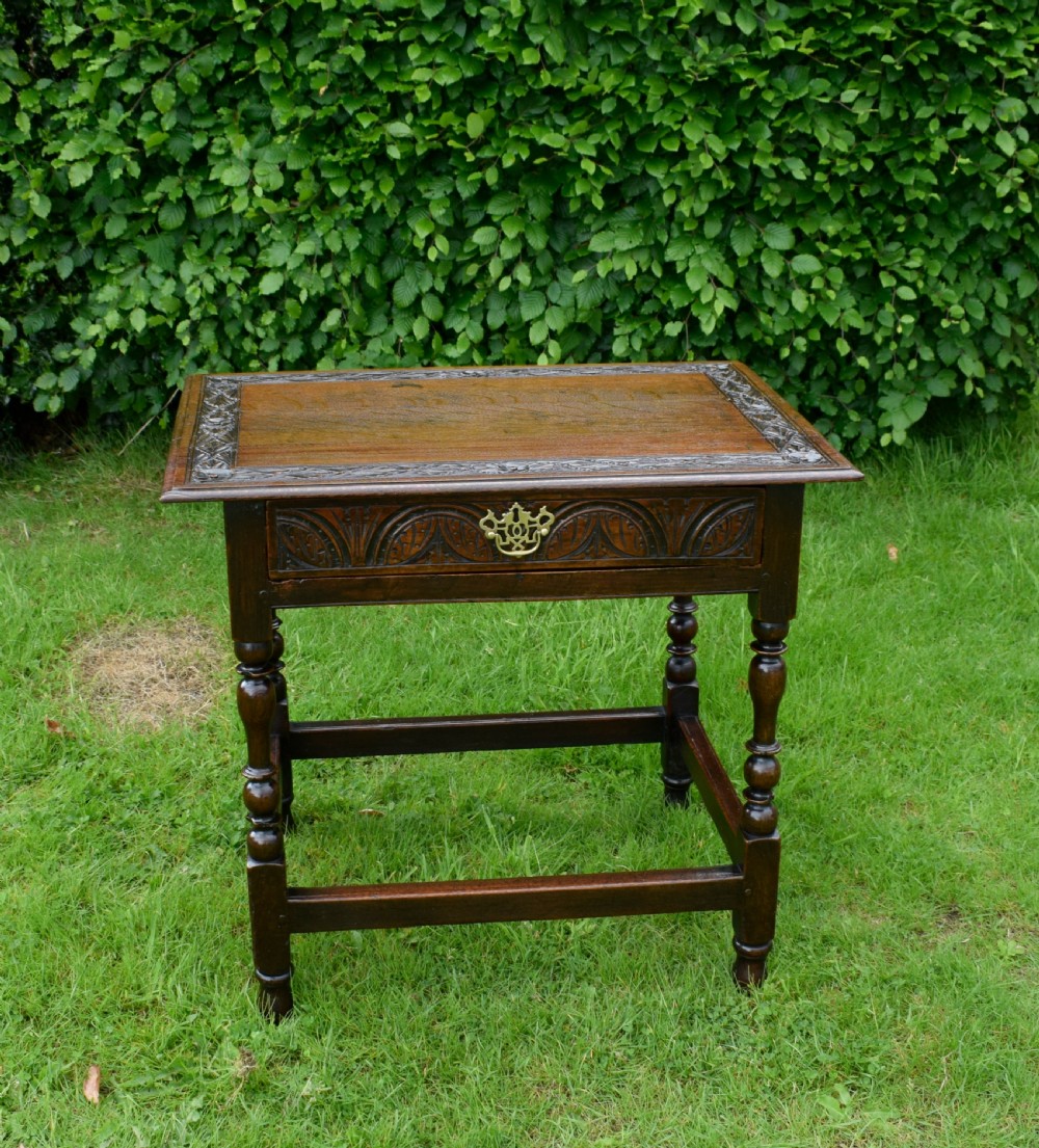 A dark oak 17th-century lowboy table featuring a carved rectangular top, a single carved drawer front with a central brass handle, turned legs, and low wooden stretchers. View V