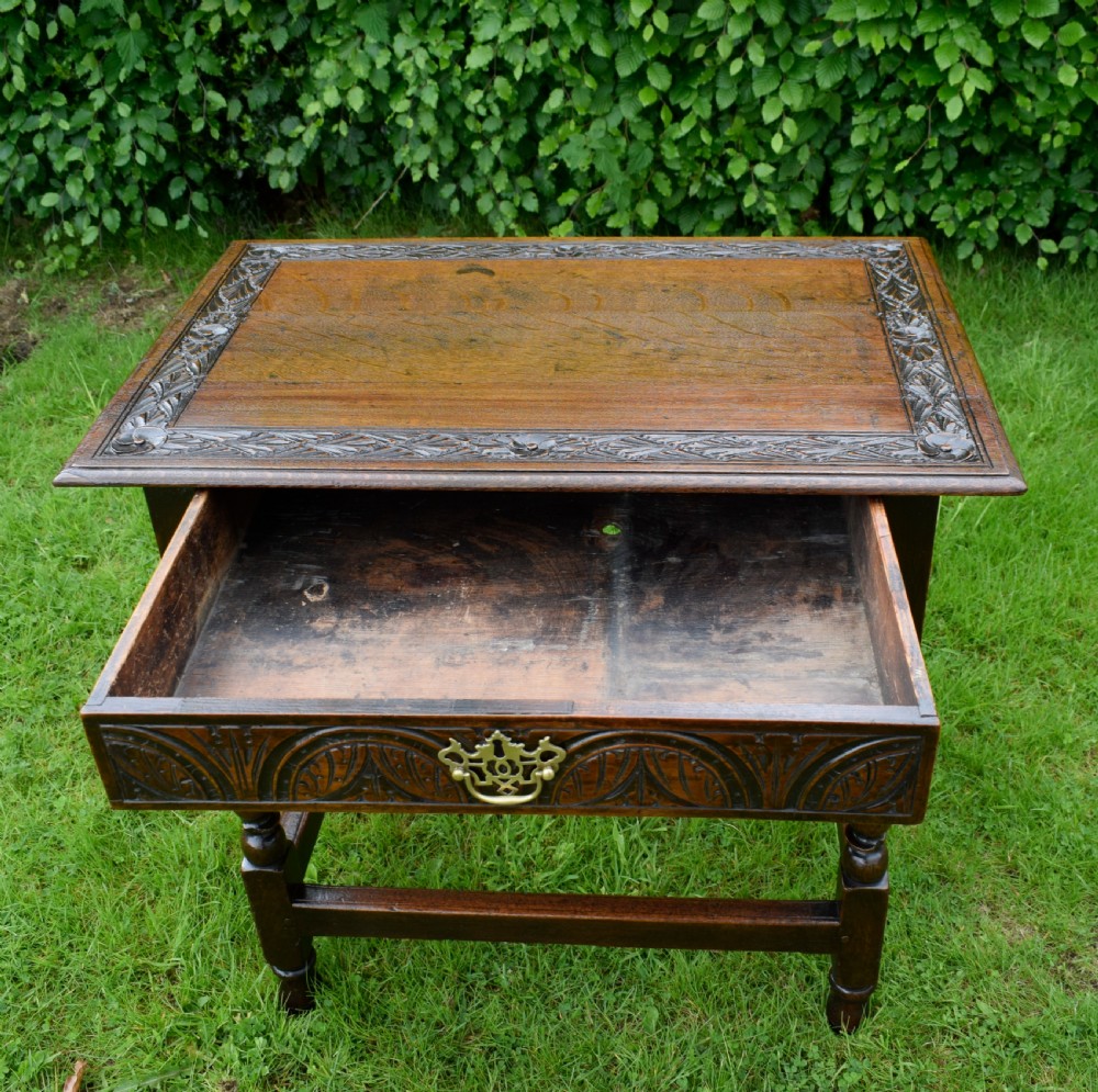 A dark oak 17th-century lowboy table featuring a carved rectangular top, a single carved drawer front with a central brass handle, turned legs, and low wooden stretchers. View VII