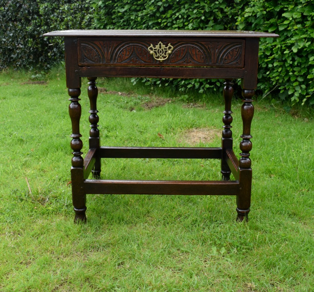 A dark oak 17th-century lowboy table featuring a carved rectangular top, a single carved drawer front with a central brass handle, turned legs, and low wooden stretchers. View VIII