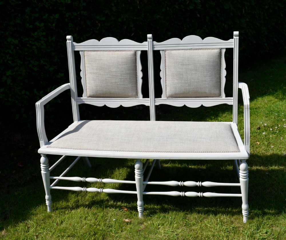A white painted Edwardian two-seater bench featuring turned legs and neutral linen upholstered seat and back panels.