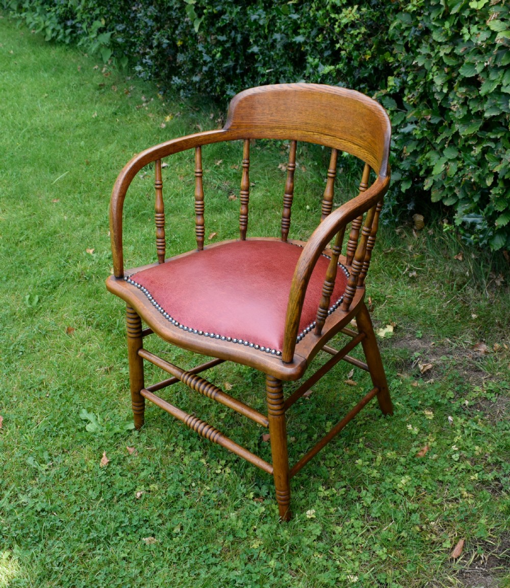 An antique oak captains chair featuring a curved horseshoe armrest, turned spindles, and a red leather upholstered seat finished with metal studding.
