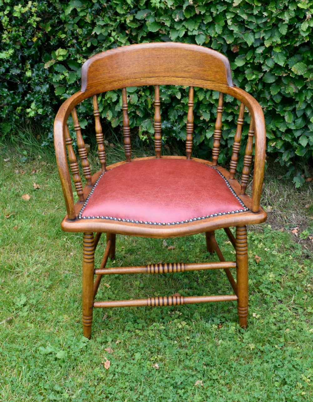 An antique oak captains chair featuring a curved horseshoe armrest, turned spindles, and a red leather upholstered seat finished with metal studding. View II