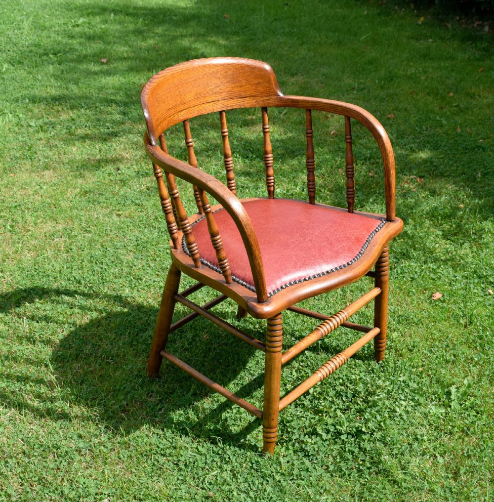 An antique oak captains chair featuring a curved horseshoe armrest, turned spindles, and a red leather upholstered seat finished with metal studding. View III