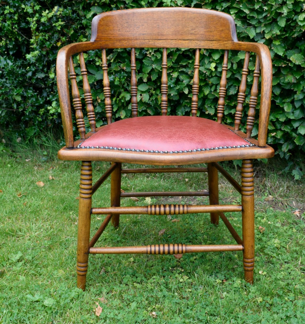 An antique oak captains chair featuring a curved horseshoe armrest, turned spindles, and a red leather upholstered seat finished with metal studding. View VI