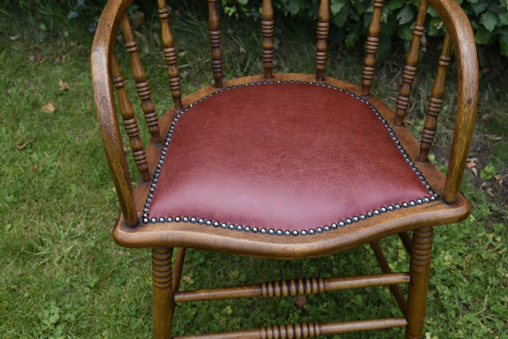 An antique oak captains chair featuring a curved horseshoe armrest, turned spindles, and a red leather upholstered seat finished with metal studding. View X