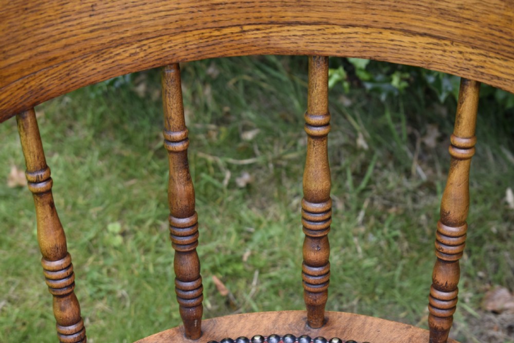 An antique oak captains chair featuring a curved horseshoe armrest, turned spindles, and a red leather upholstered seat finished with metal studding. View XII
