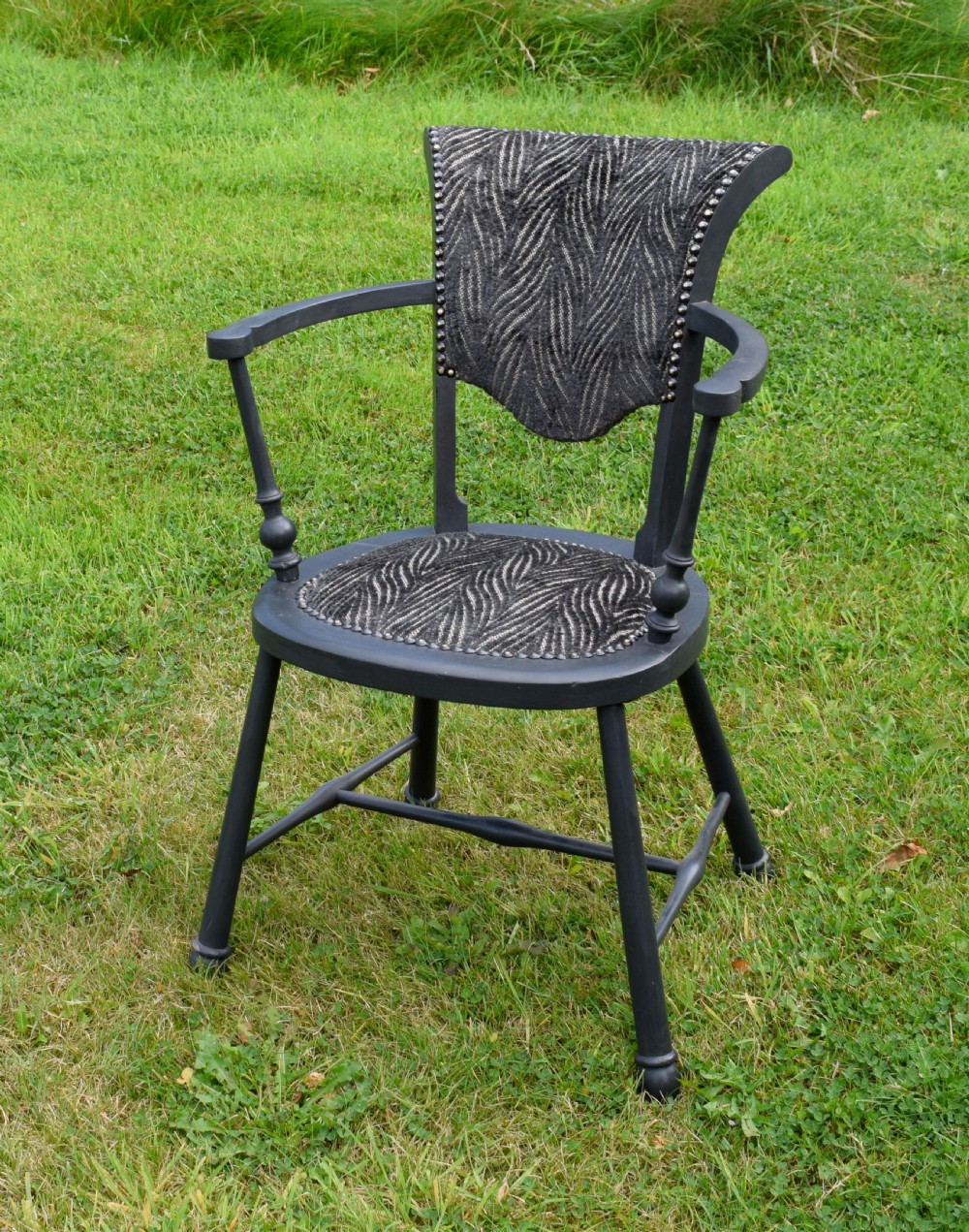 A dark grey painted Arts and Crafts style elbow chair featuring textured patterned fabric upholstery on the seat and curved backrest.