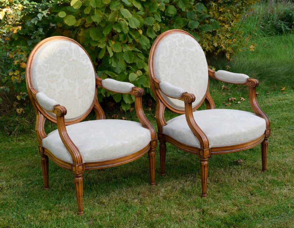 A pair of French Louis XVI style open armchairs featuring carved wooden frames, oval medallion backs, fluted legs, and cream damask upholstery.