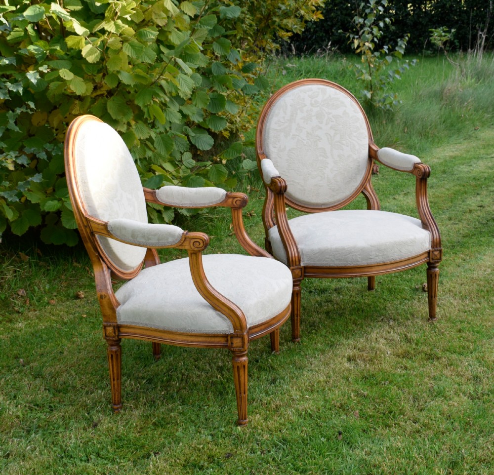 A pair of French Louis XVI style open armchairs featuring carved wooden frames, oval medallion backs, fluted legs, and cream damask upholstery. View II