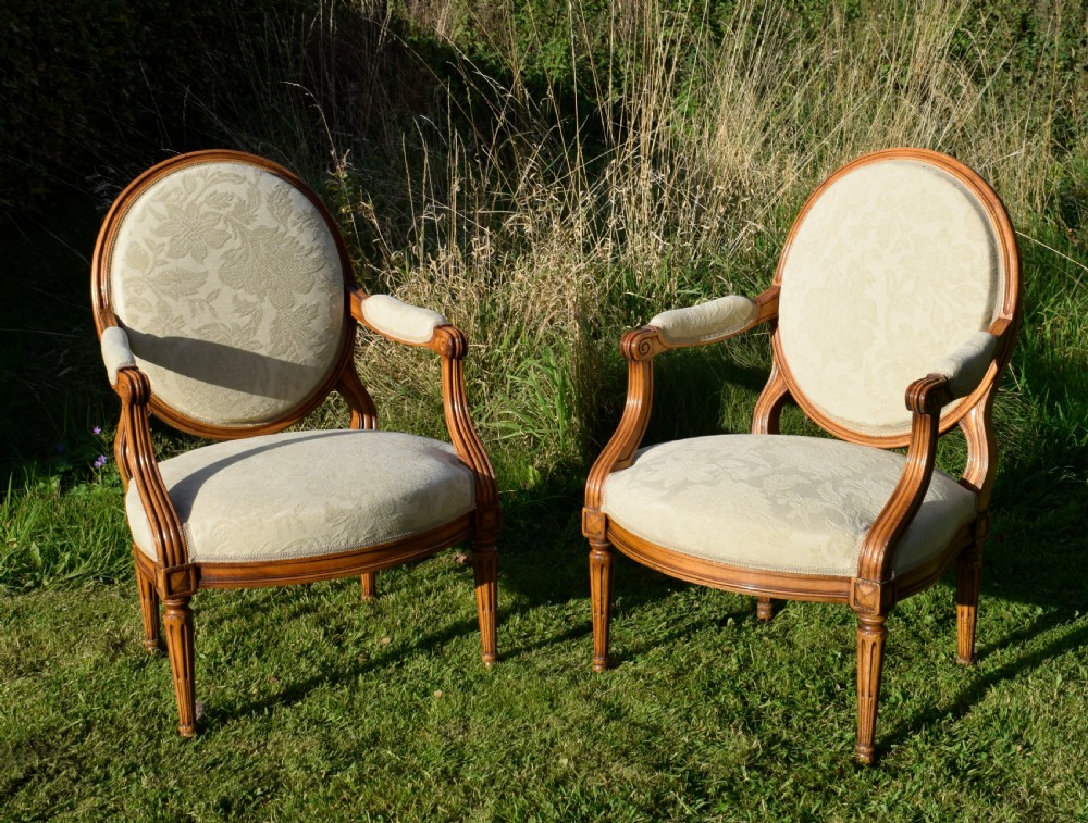 A pair of French Louis XVI style open armchairs featuring carved wooden frames, oval medallion backs, fluted legs, and cream damask upholstery. View III