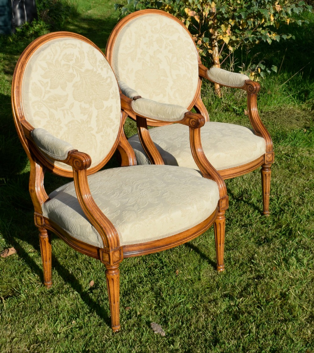 A pair of French Louis XVI style open armchairs featuring carved wooden frames, oval medallion backs, fluted legs, and cream damask upholstery. View V