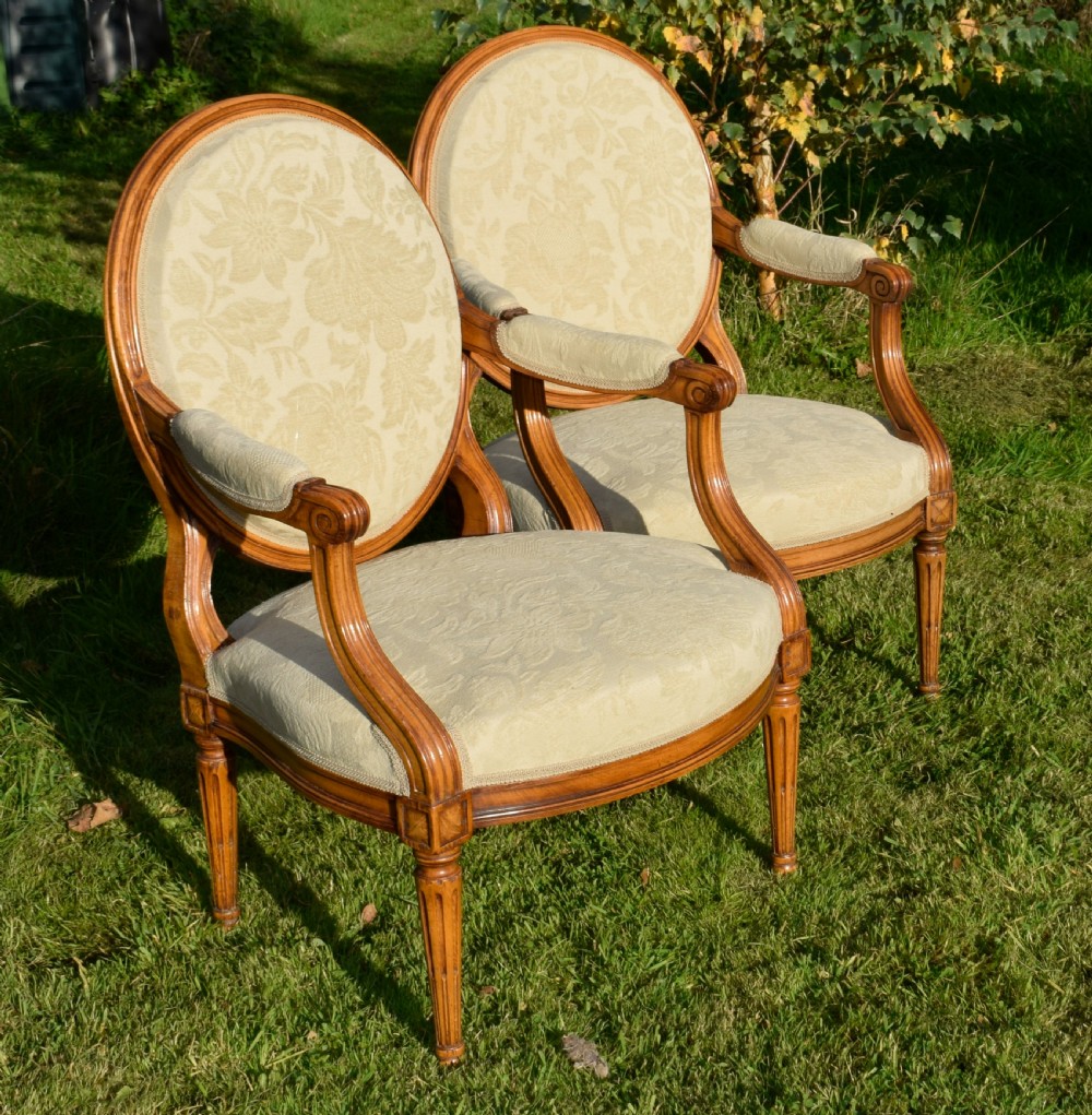 A pair of French Louis XVI style open armchairs featuring carved wooden frames, oval medallion backs, fluted legs, and cream damask upholstery. View VI