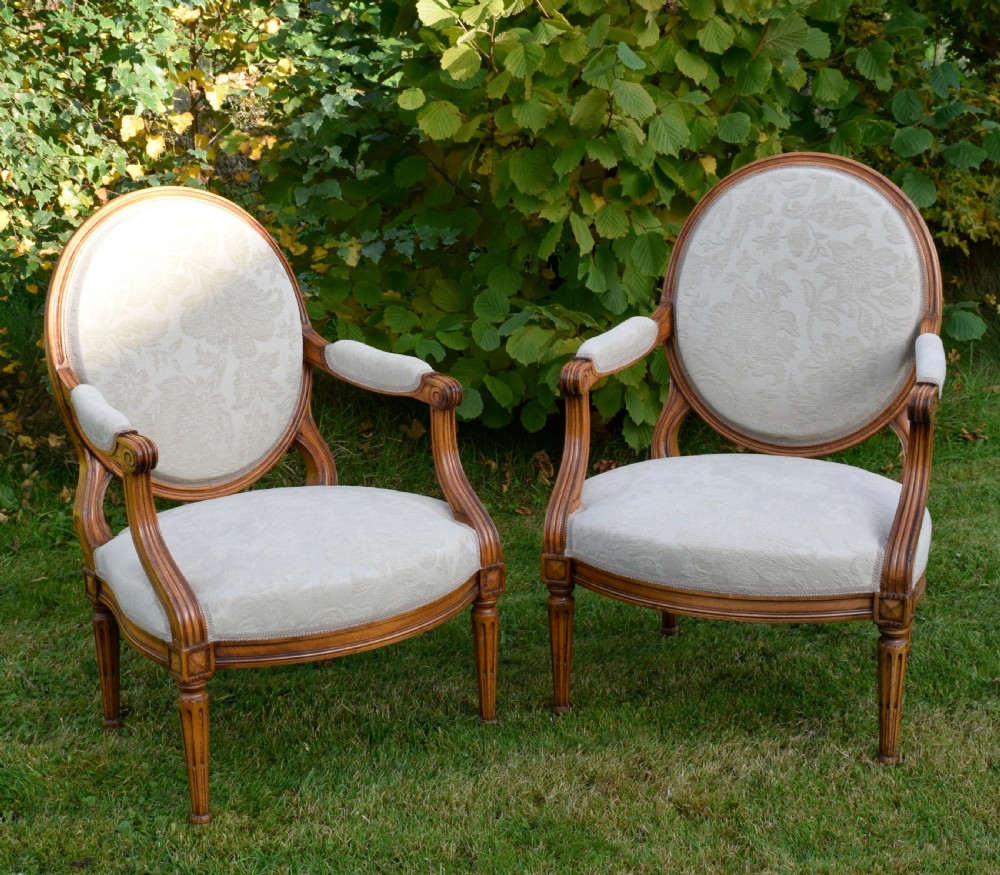 A pair of French Louis XVI style open armchairs featuring carved wooden frames, oval medallion backs, fluted legs, and cream damask upholstery. View X