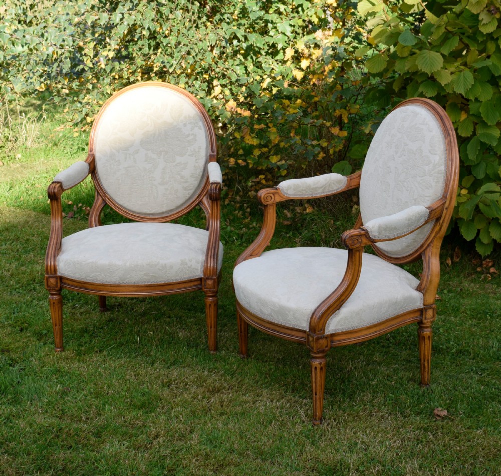 A pair of French Louis XVI style open armchairs featuring carved wooden frames, oval medallion backs, fluted legs, and cream damask upholstery. View XIII