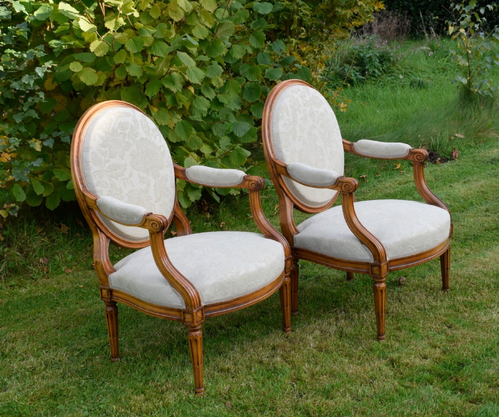 A pair of French Louis XVI style open armchairs featuring carved wooden frames, oval medallion backs, fluted legs, and cream damask upholstery. View XV