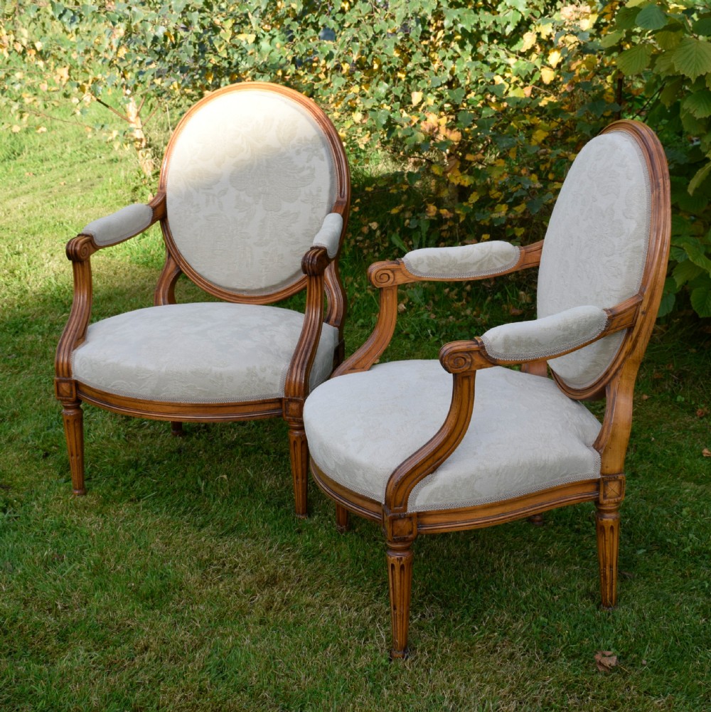 A pair of French Louis XVI style open armchairs featuring carved wooden frames, oval medallion backs, fluted legs, and cream damask upholstery. View XVI