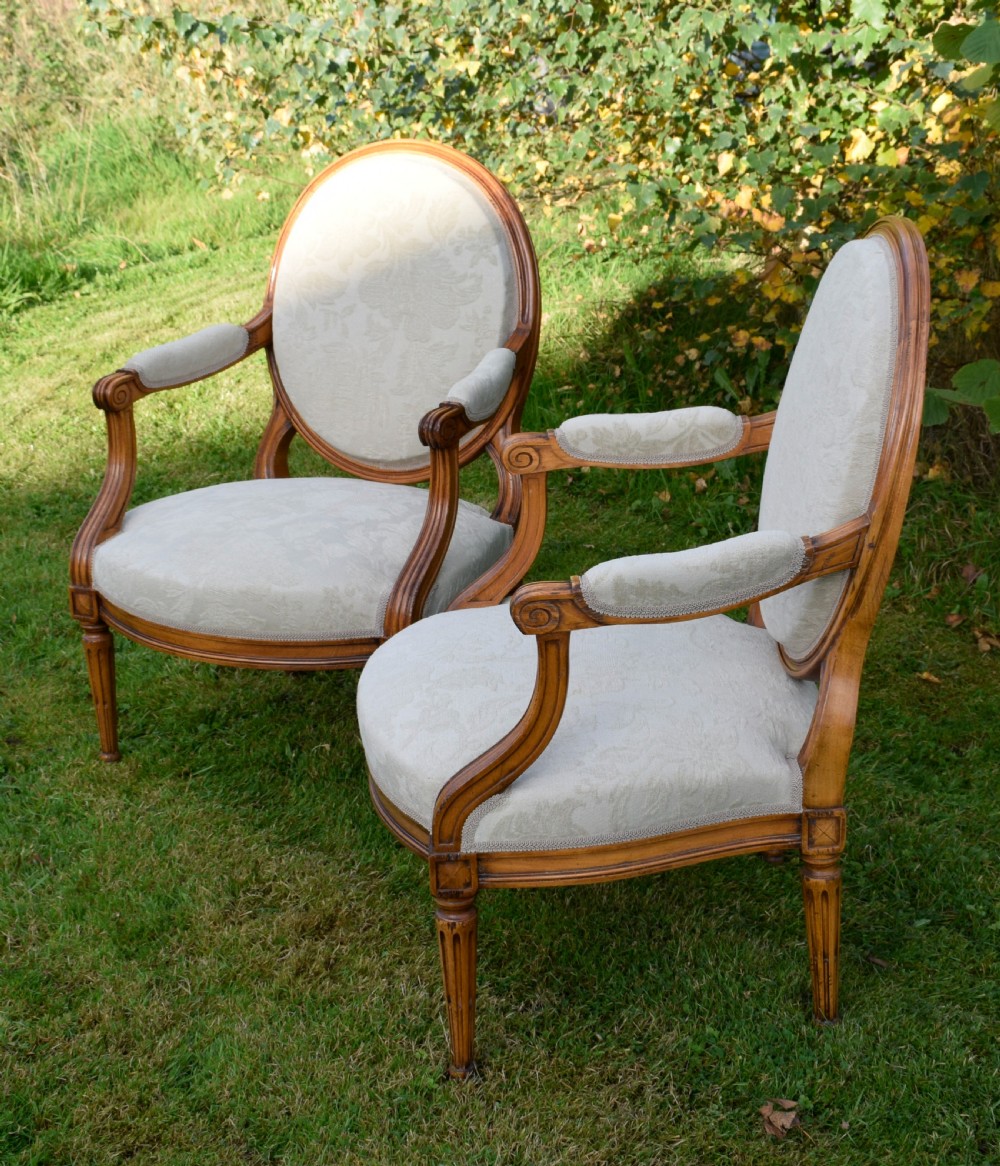 A pair of French Louis XVI style open armchairs featuring carved wooden frames, oval medallion backs, fluted legs, and cream damask upholstery. View XVII