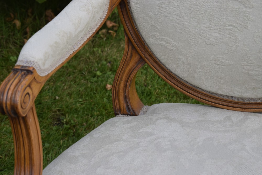 A pair of French Louis XVI style open armchairs featuring carved wooden frames, oval medallion backs, fluted legs, and cream damask upholstery. View XVIII