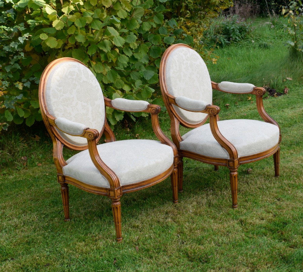 A pair of French Louis XVI style open armchairs featuring carved wooden frames, oval medallion backs, fluted legs, and cream damask upholstery. View XX