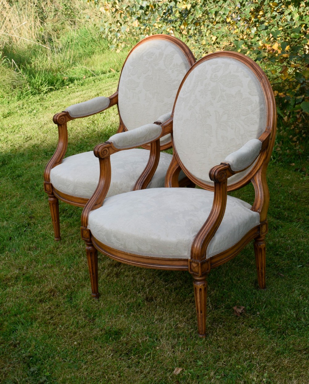 A pair of French Louis XVI style open armchairs featuring carved wooden frames, oval medallion backs, fluted legs, and cream damask upholstery. View XXI