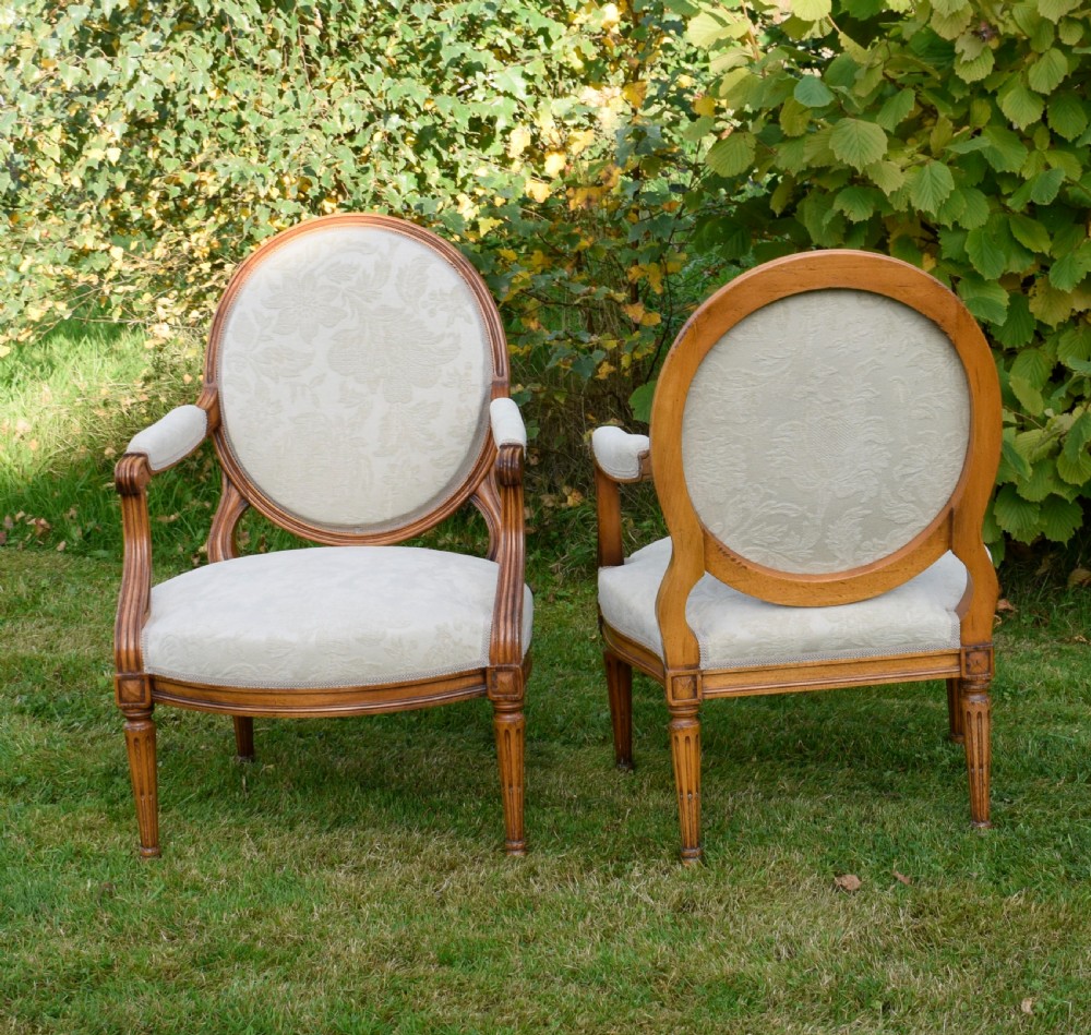 A pair of French Louis XVI style open armchairs featuring carved wooden frames, oval medallion backs, fluted legs, and cream damask upholstery. View XXII