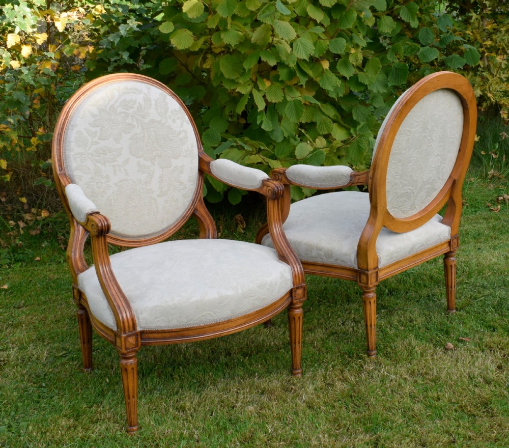 A pair of French Louis XVI style open armchairs featuring carved wooden frames, oval medallion backs, fluted legs, and cream damask upholstery. View XXIII