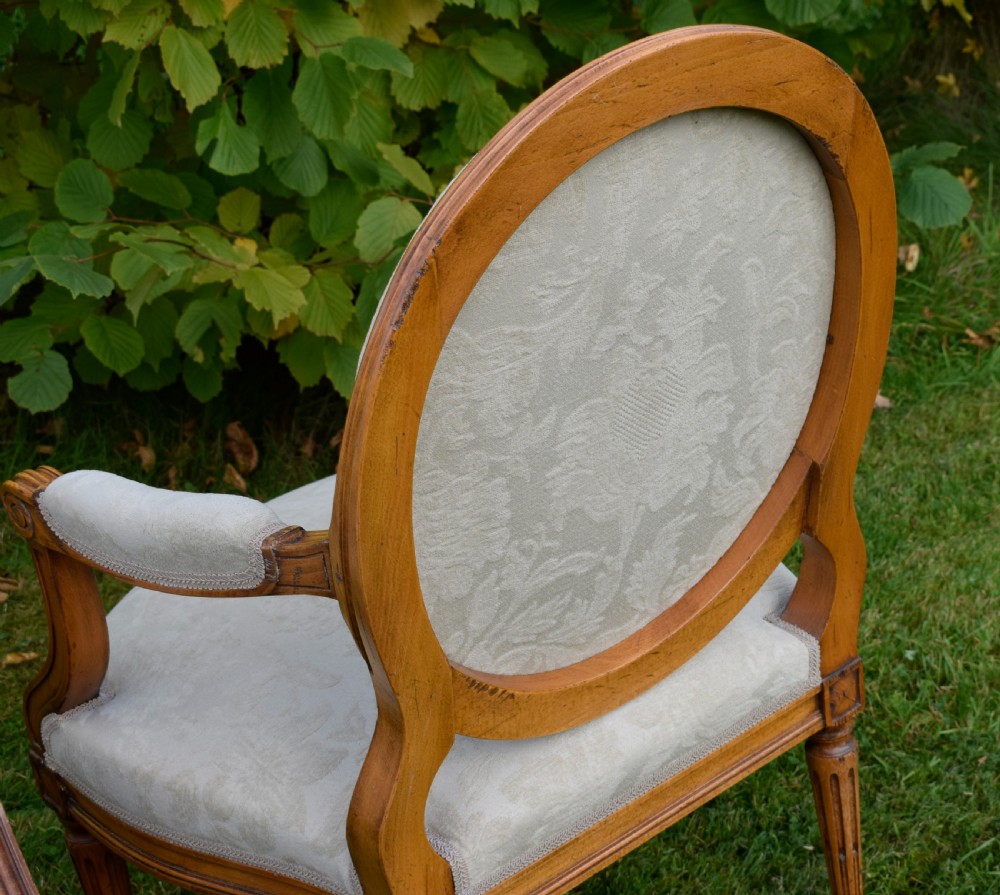 A pair of French Louis XVI style open armchairs featuring carved wooden frames, oval medallion backs, fluted legs, and cream damask upholstery. View XXV