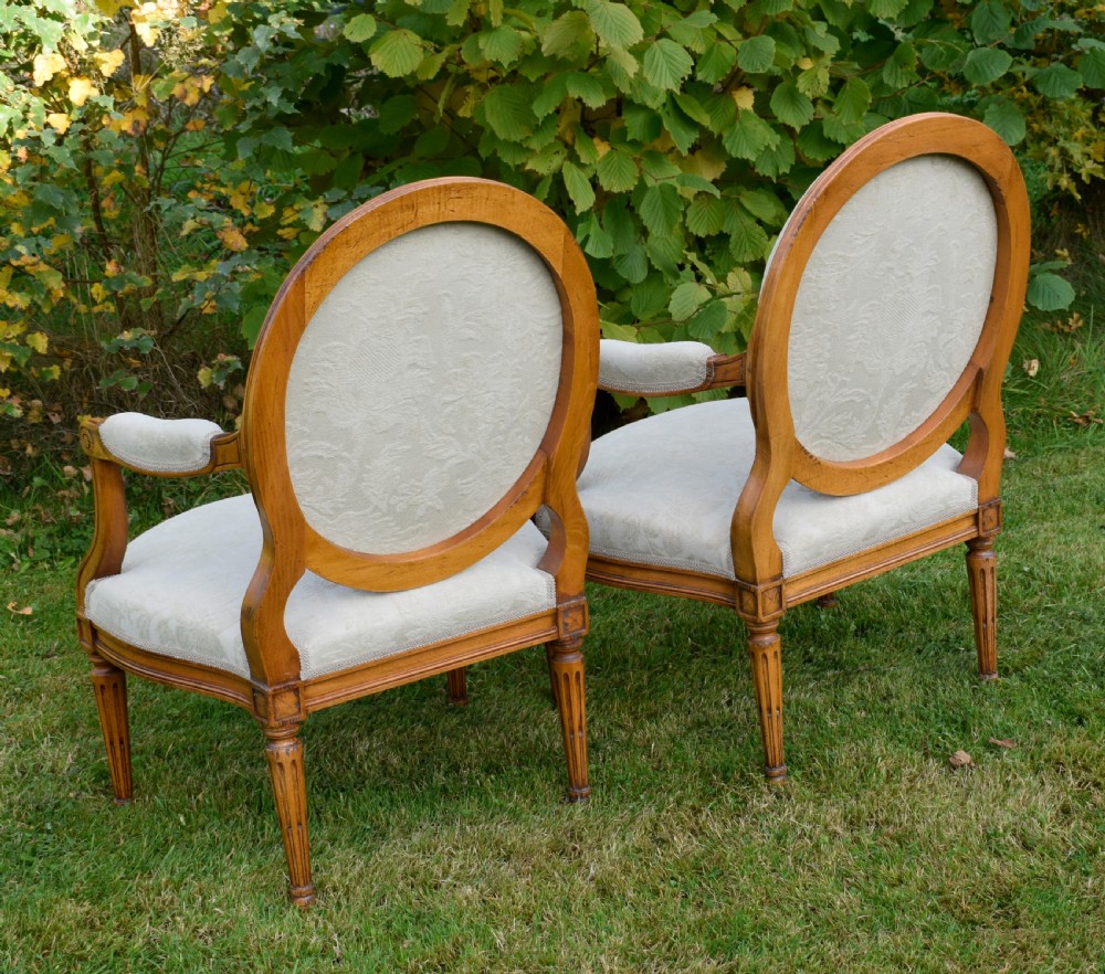 A pair of French Louis XVI style open armchairs featuring carved wooden frames, oval medallion backs, fluted legs, and cream damask upholstery. View XXVI