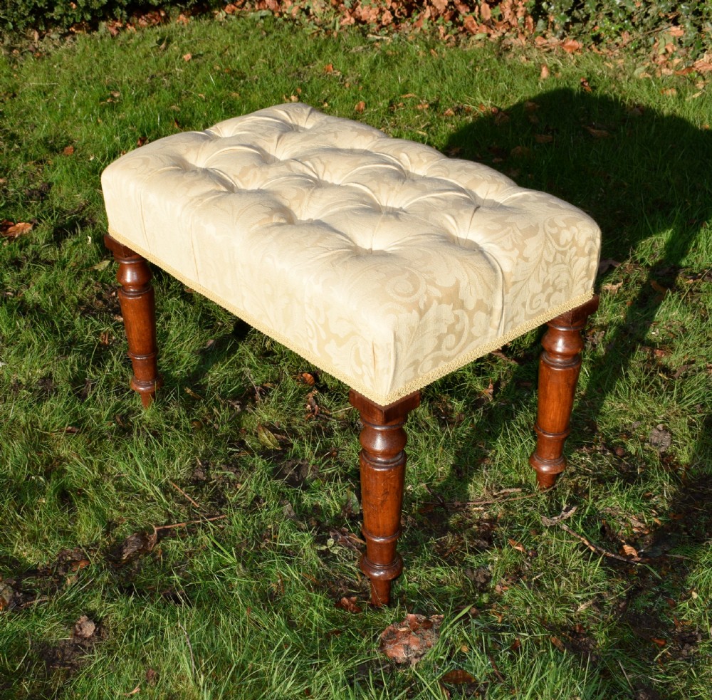 A Victorian upholstered rectangular footstool featuring cream damask fabric with deep buttoning, braid trim, and four dark turned wooden legs.
