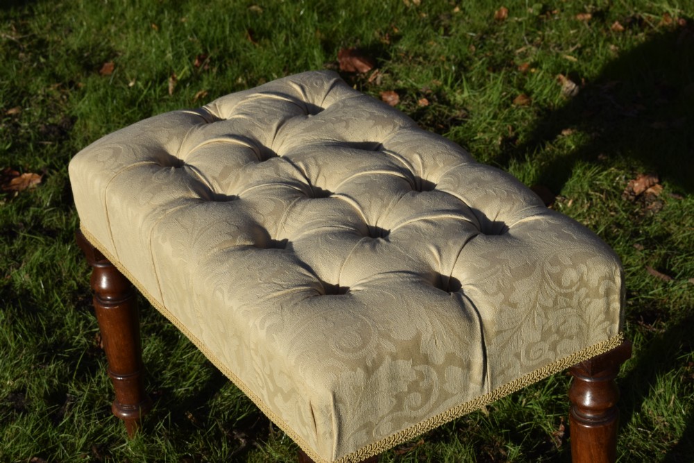 A Victorian upholstered rectangular footstool featuring cream damask fabric with deep buttoning, braid trim, and four dark turned wooden legs. View VI