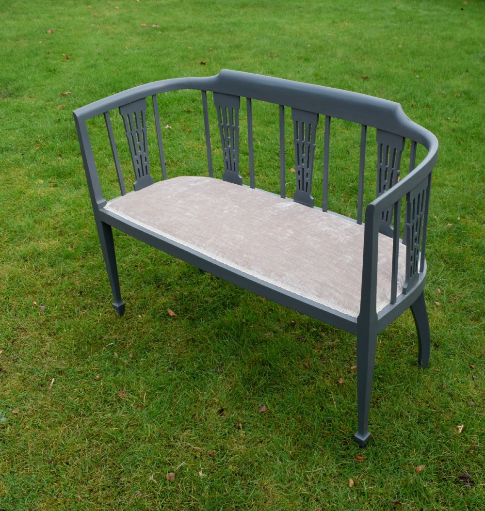 An Edwardian painted grey wooden settee featuring vertical spindles, pierced back splats, and a neutral beige velvet upholstered seat.