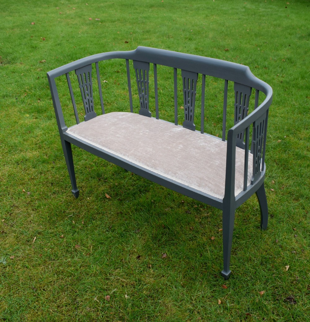 An Edwardian painted grey wooden settee featuring vertical spindles, pierced back splats, and a neutral beige velvet upholstered seat. View II