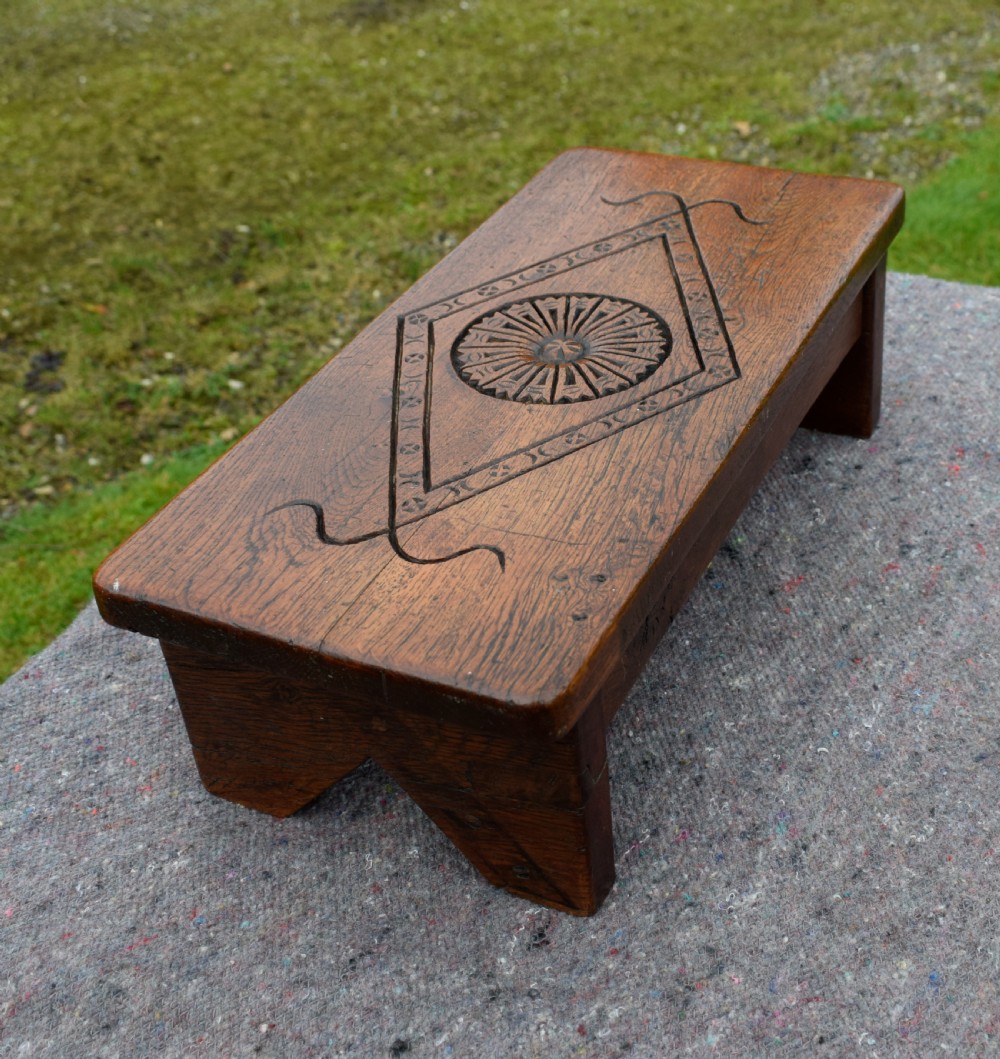 A rectangular warm brown carved oak footstool featuring a central rosette enclosed in a lozenge motif set upon slab legs.