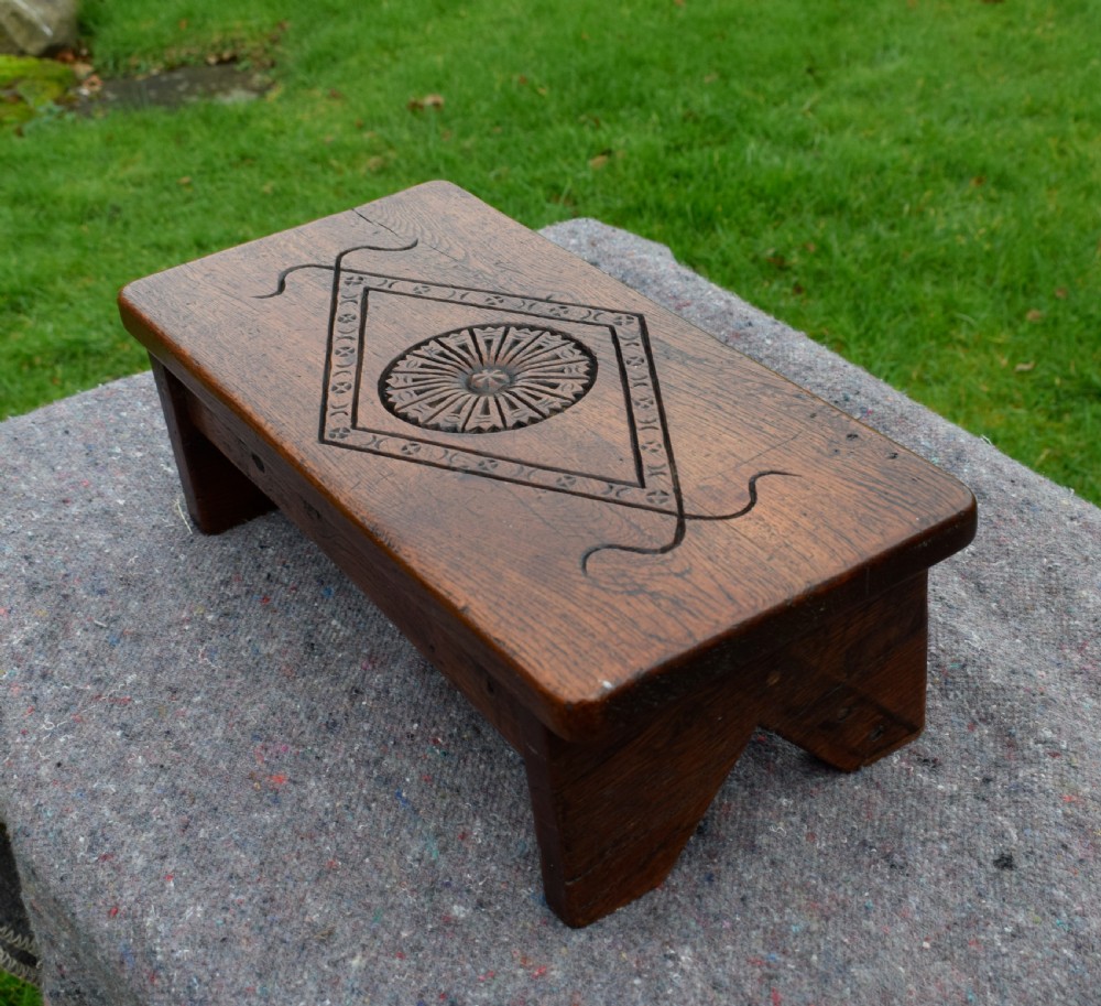 A rectangular warm brown carved oak footstool featuring a central rosette enclosed in a lozenge motif set upon slab legs. View II