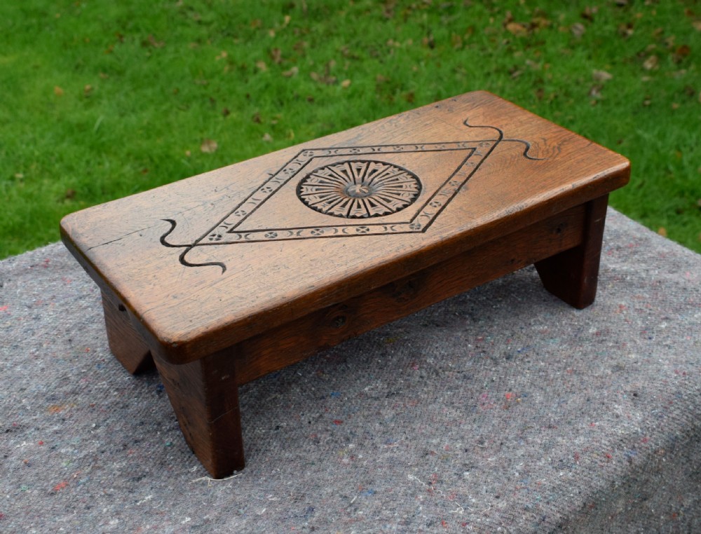 A rectangular warm brown carved oak footstool featuring a central rosette enclosed in a lozenge motif set upon slab legs. View IV