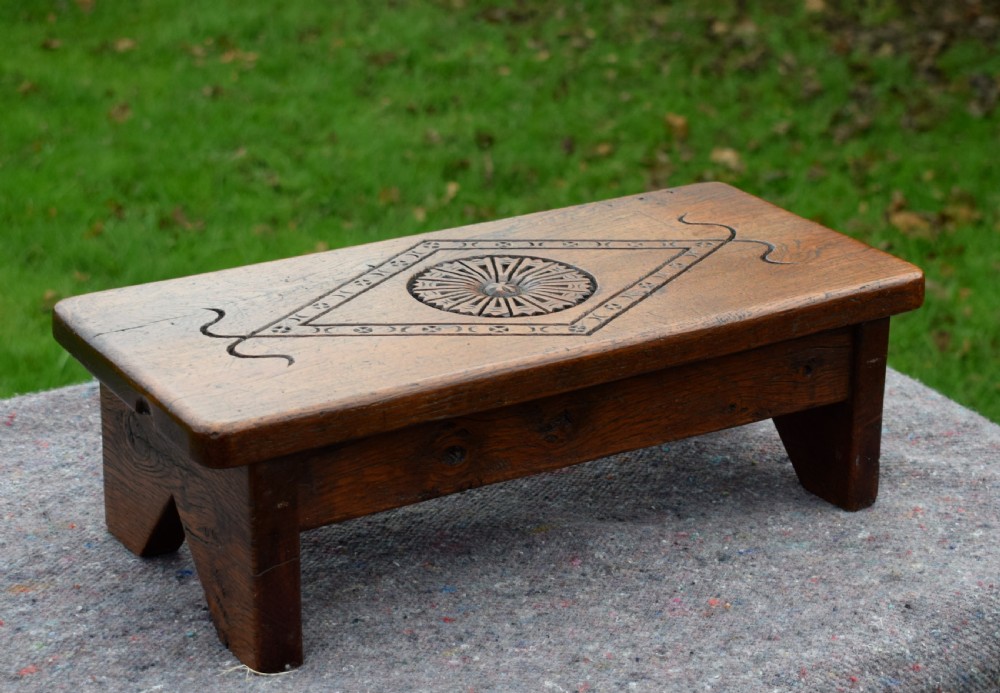 A rectangular warm brown carved oak footstool featuring a central rosette enclosed in a lozenge motif set upon slab legs. View VII