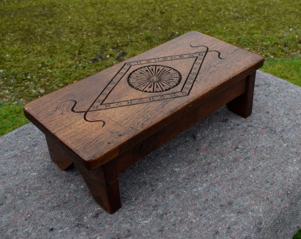 A rectangular warm brown carved oak footstool featuring a central rosette enclosed in a lozenge motif set upon slab legs. View IX
