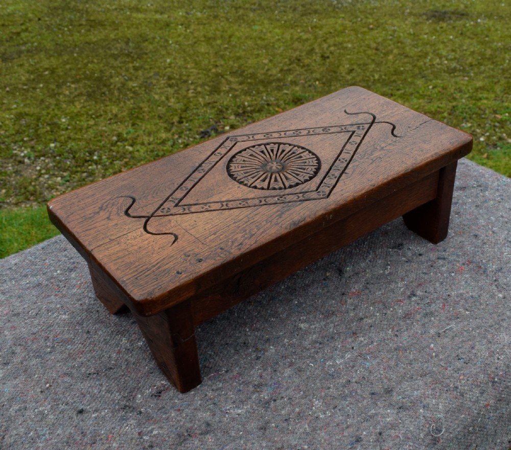 A rectangular warm brown carved oak footstool featuring a central rosette enclosed in a lozenge motif set upon slab legs. View X